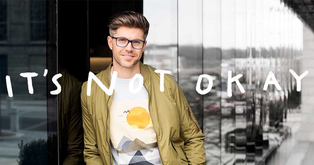 Darren Kennedy leaning against a glass wall with the words 'It's not okay' overlaid