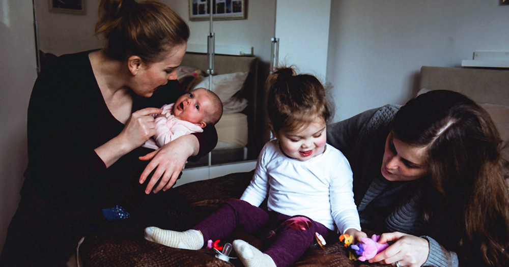 Two women at home with their daughters, one a baby in arms and the other a toddler playing on the couch, one of the many same-sex families in Ireland