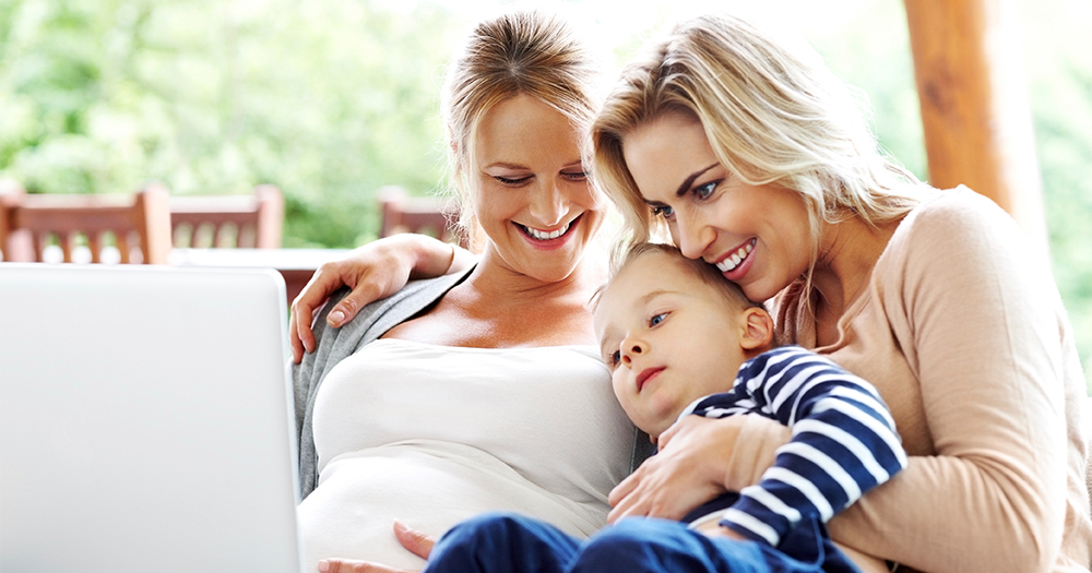 A family with two mothers, one pregnant, and their little boy all smiling on the couch looking at a laptop