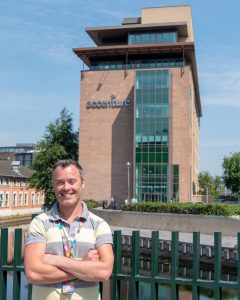 A man stands outside a building smiling. It's a sunny day.