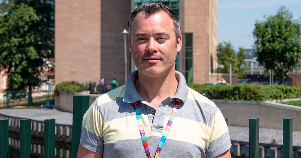 A man stands outside a building smiling. It's a sunny day.