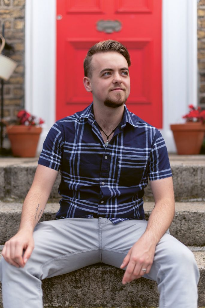 Man sitting on steps. Behind him a red door: :in this piece queer people describe the difficulties of a simple trip to the barbershop