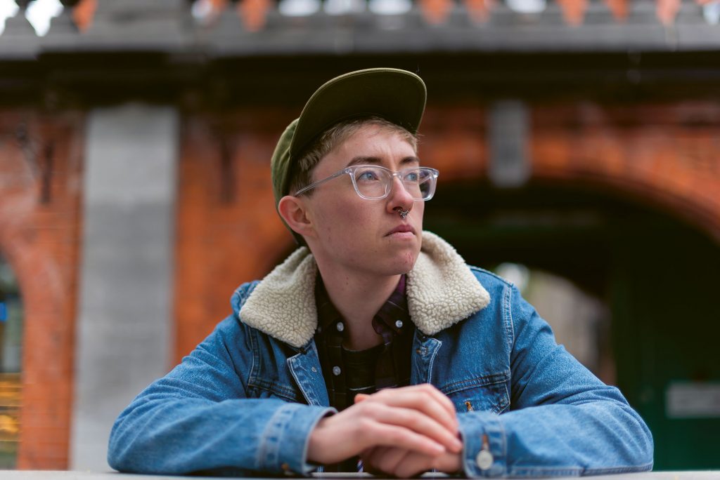 Non binary person wearing a baseball cap and glasses: in this piece queer people describe the difficulties of a simple trip to the barbershop