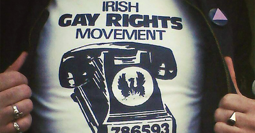 A man pulling back his leather jacket to reveal a t-shirt reading Irish Gay Rights Movement and a picture of a telephone