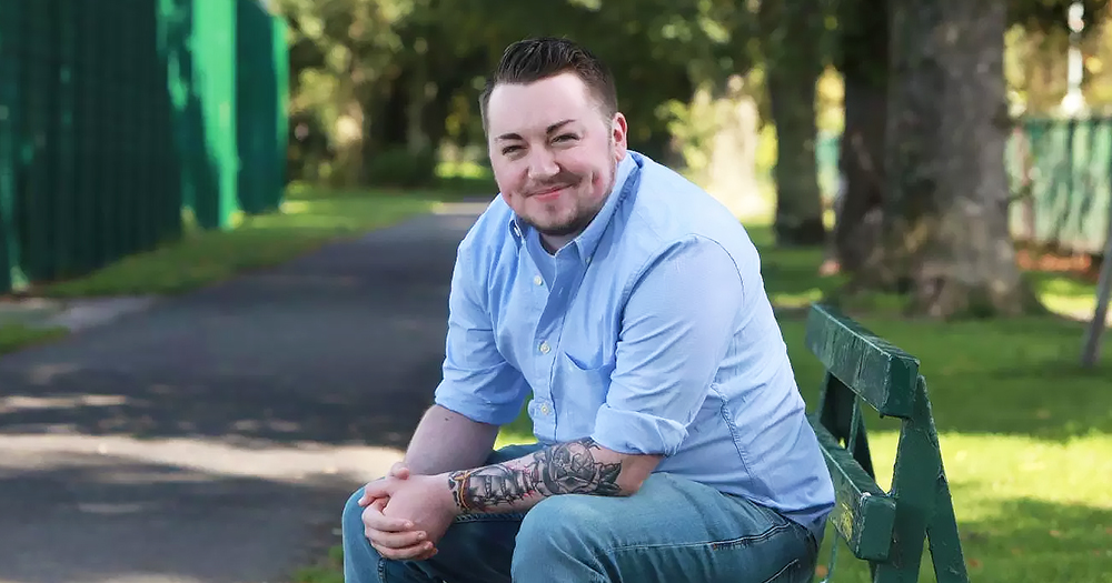 Jack Murphy, a young trans man with a beard, smiles at the camera while sitting on a park bench