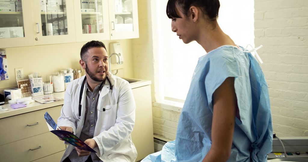Doctor talking to patient wearing a hospital gown sitting on a bed. A new study on trans healthcare has been launched.