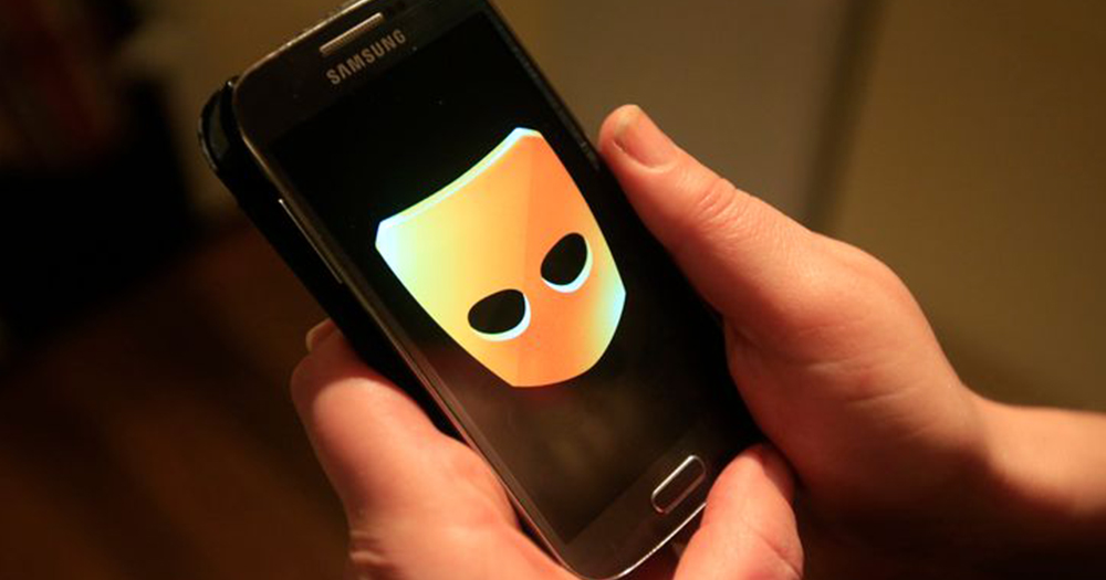 Close up of two hands holding a Samsung phone with the Grindr logo, a yellow mask with black eyeholes, coming up on the screen. A teenage boy was kidnapped after he met someone on the dating app.