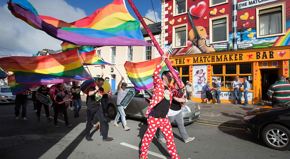 A number of people dressed in brightly coloured clothes are walking down a road waving rainbow flags. They are walking past a pub called tThe Matchmaker which has a yellow sign and is adorned with red hearts.