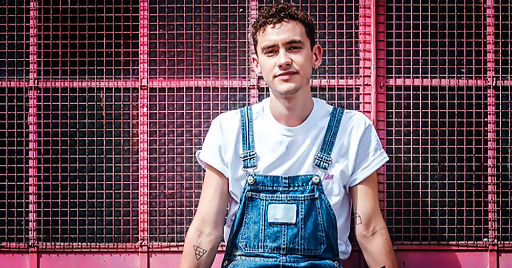 Singer Olly Alexander wearing a white tshirt and a dungaree, leaning against a pink gate