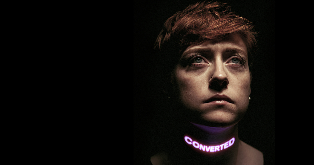 A woman in the shadows with the word Converted in lights across her neck
