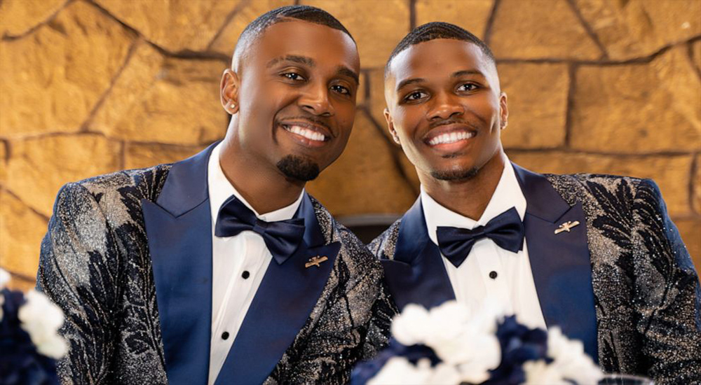 The picture shows two grooms on their wedding day. They are wearing flowery blue suits and there is a yellow background. They look really happy.