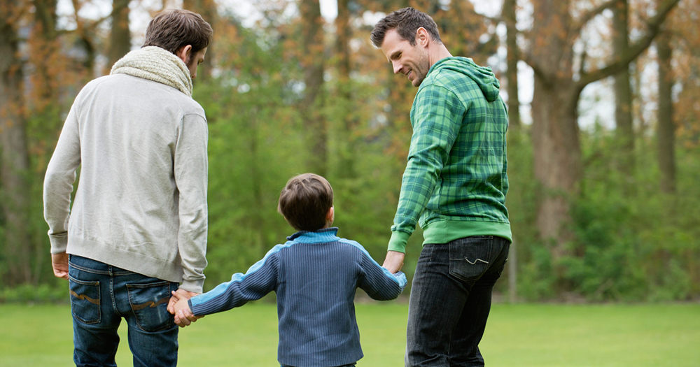 Two men hold the hands of a child as they enjoy a family walk in the woods. The are wearing green, blue and white sweaters repectively.