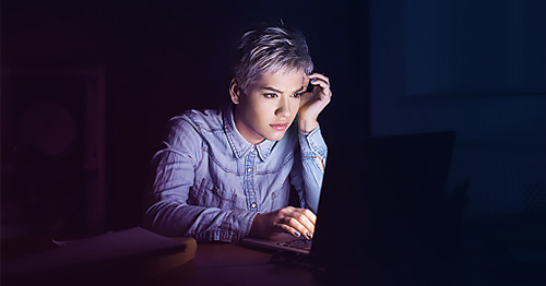 Woman with short hair and a denim shirt looking at digital content on her laptop in a dark room.