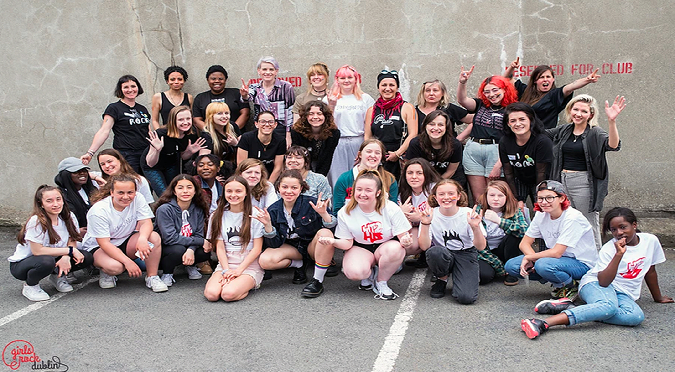 A group of teenage girls and non-binary people sitting or standing in front of a grey wall wearing Girls Rock Dublin t-shirts and looking happy.