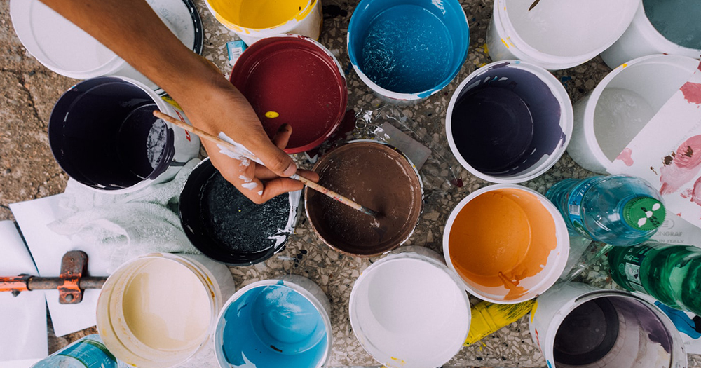 Hand holding paintbrush dipped in paint surrounded by other pots of paint