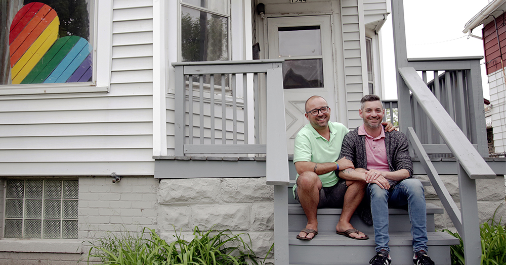 A smiling gay couple sit on the outdoor steps of a house with a rainbow heart in the window