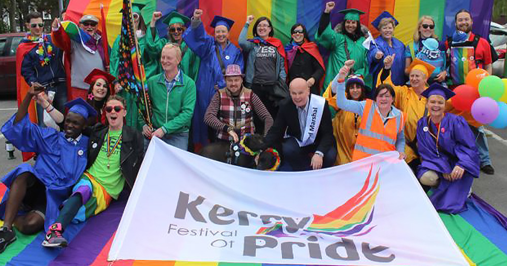 The Kerry Pride team all in rainbow colours with a huge rainbow flag behind them, holding a huge banner
