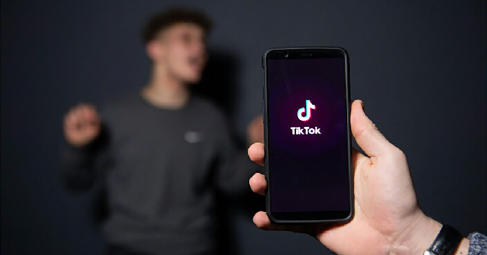 A hand holding a phone with TikTok logo on it, in the background a man out of focus appears to be singing