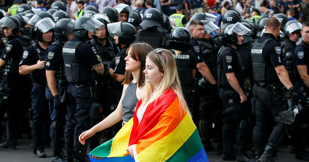 Two women walking in front of police officers, one of them wearing a rainbow flag. A bisexual woman has called for an investigation into torture in Chechnya.