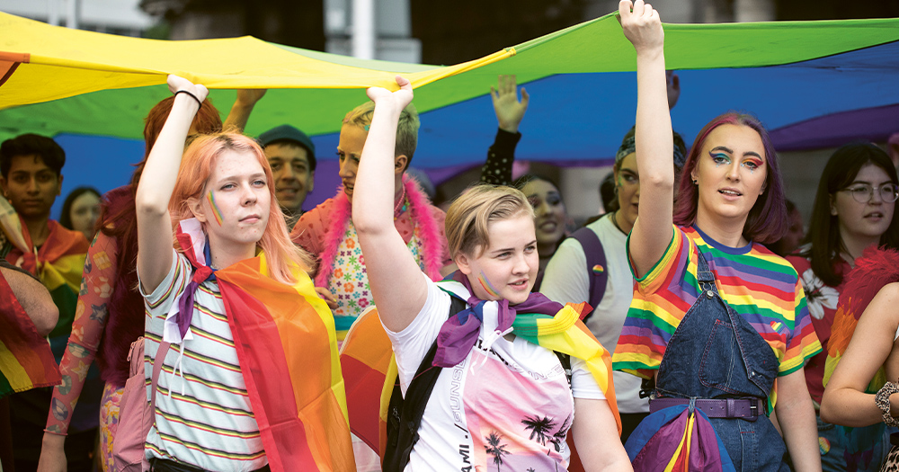 A group of young people at a Pride parade holding a huge rainbow flag over their head