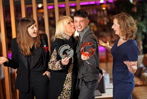 Anne Doyle kisses James Kavanag on the cheek while they hold awards, two amused women looking on