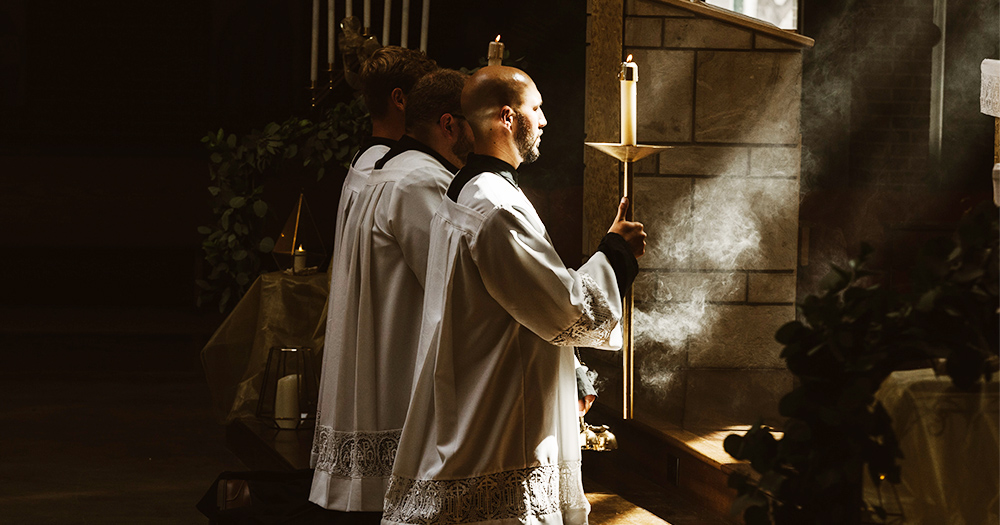 Three priests kneel in a shaft of sunlight, holding candles