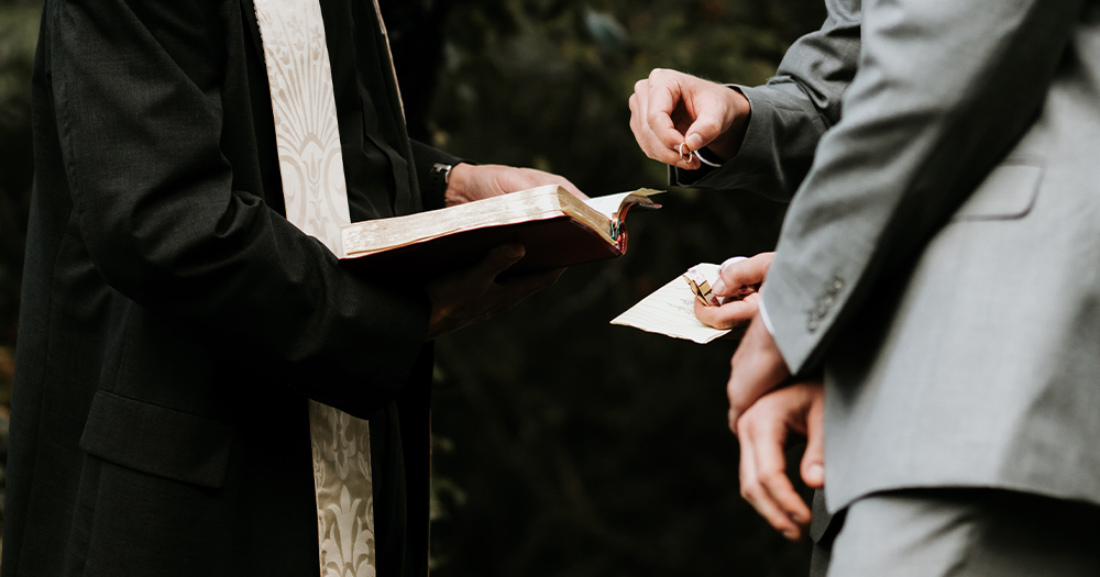 A priest performs a marriage ceremony and two men in grey suits stand before him holding a ring. none of their heads are visible