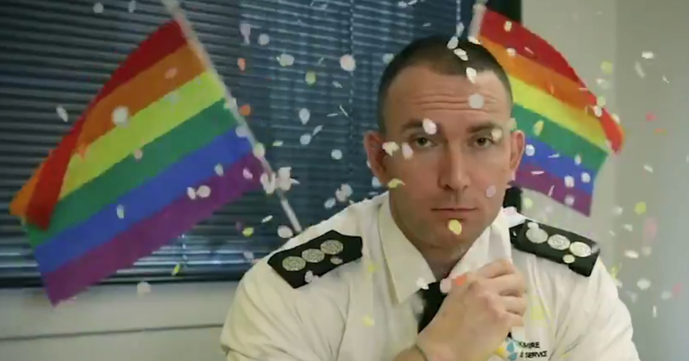 Firefighter sits at table while LGBT+ rainbow flags wave in the background along with confetti.