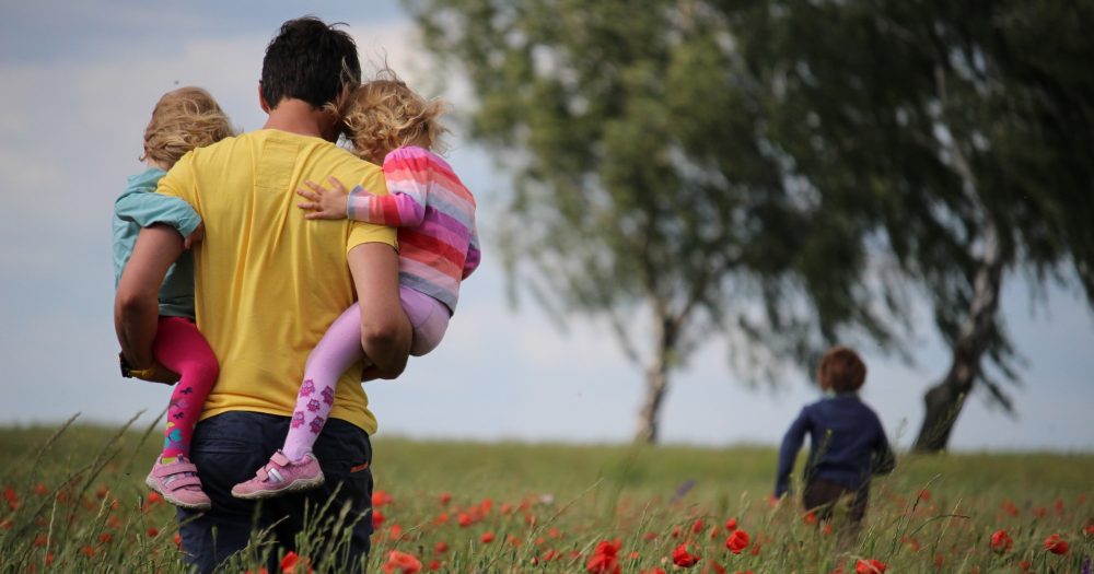 queer dad surrogacy Man carries his two daughters through field. His sun is running in the background. gay dad, surrogacy