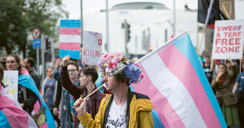 A Trans Pride march with a young man in the foreground with flowers on his head carrying a huge trans flag