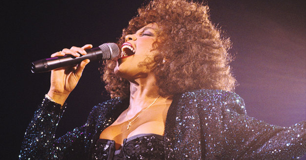 Whitney Houston signing into a microphone wearing a black sparkling jacket and dress, her songs will be played during Whitney Houston: Hologram Tour