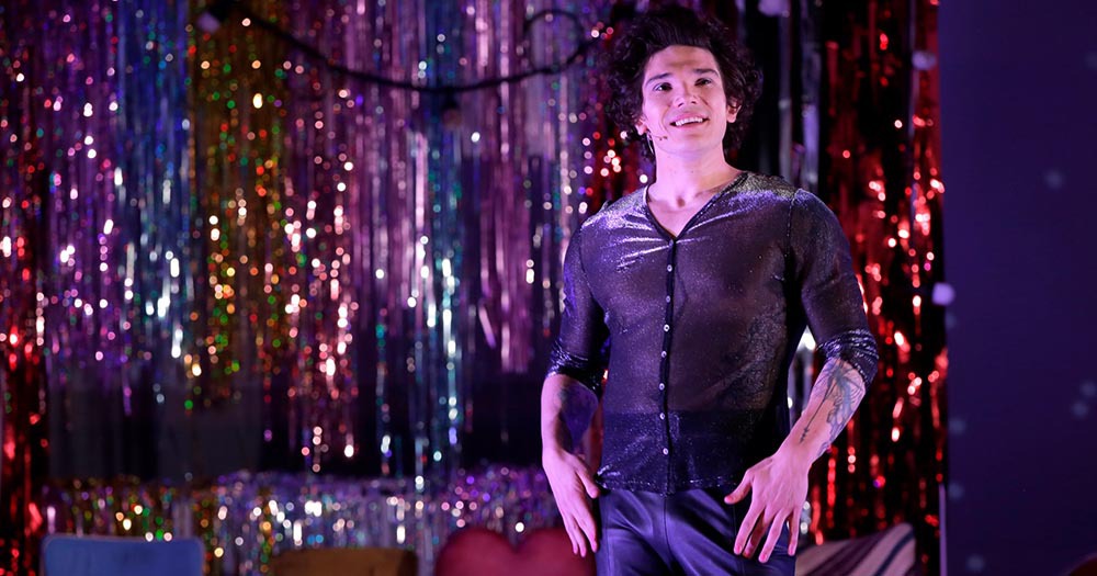 Cis man, Fra Fee, wearing a shimmering shirt and standing in front of rainbow tinsel for the role of trans woman Pussy in Breakfast on Pluto musical