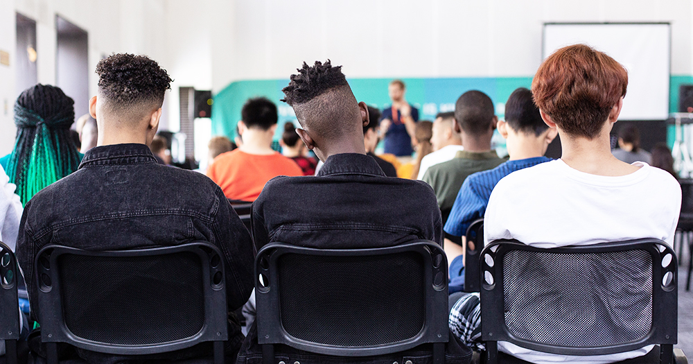 Seen from behind, a group of young people seated in a class are looking at a lecturer