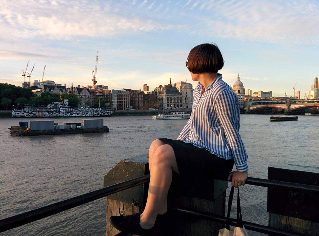 Russian woman sitting on a fence in front of a river looking at the city in front of her and ship in the river