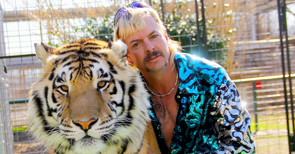 A flamboyantly dressed man with bleached hair and a moustache embraces a tiger inside a cage