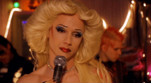 A drag artist with 1970's style hair holds a microphone and looks wistfully into the distance