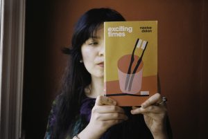 A woman with long dark hair holds a book up to cover half of her face