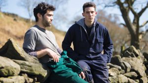 Two men dressed in work clothes lean against a stone wall in the countryside