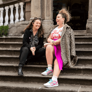 Two laughing hipster women sits on the steps leading up to a building