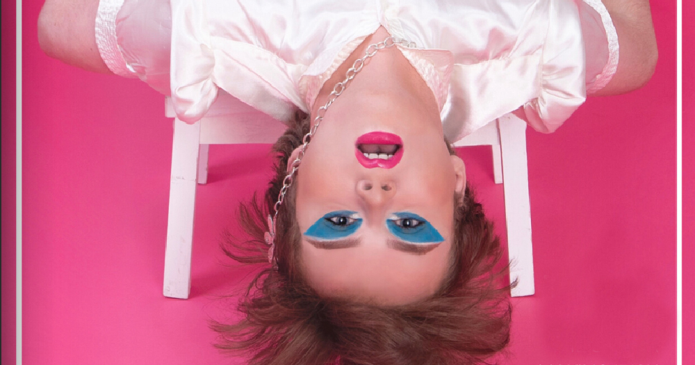 A young man wearing makeup hangs upside down against a pink background