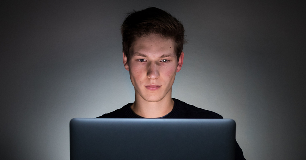 A young man in a dark room, his face lit up by the glow of a laptop screen