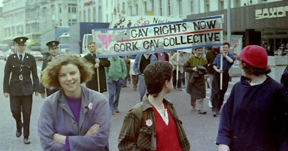 An old photo of the Cork Gay Collective holding a banner walking down a street accompanied by policemen while three women look on
