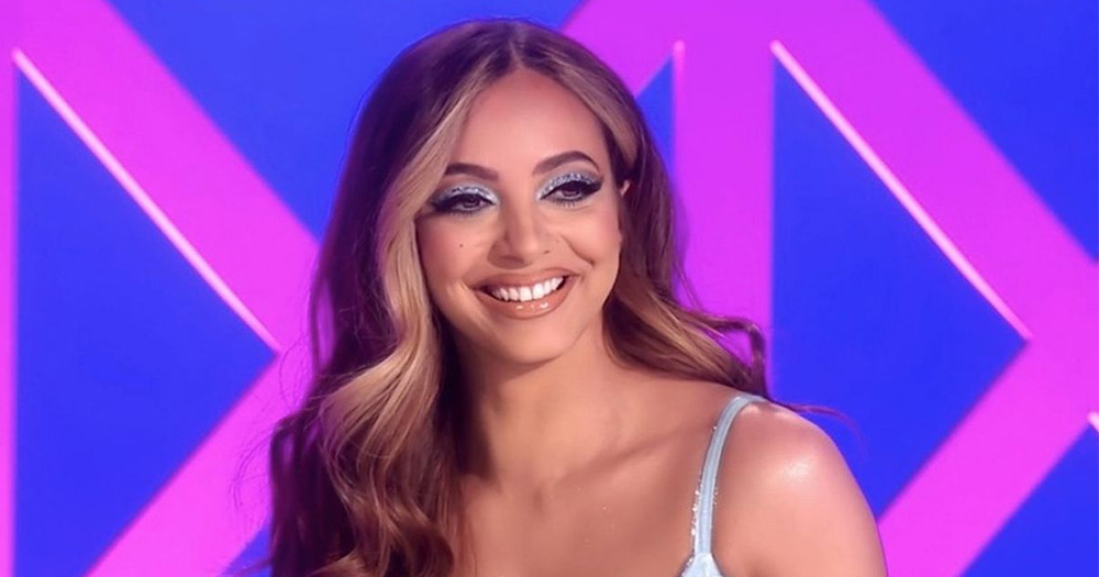 Jade from Little Mix with long wavy hair and strappy dress smiling in front of a multicoloured background