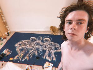 A topless longhaired young man takes a selfie with a painting his creating lying on the floor behind him