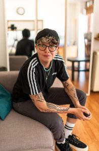 A woman wearing a cap with lots of tattoos sits on a couch