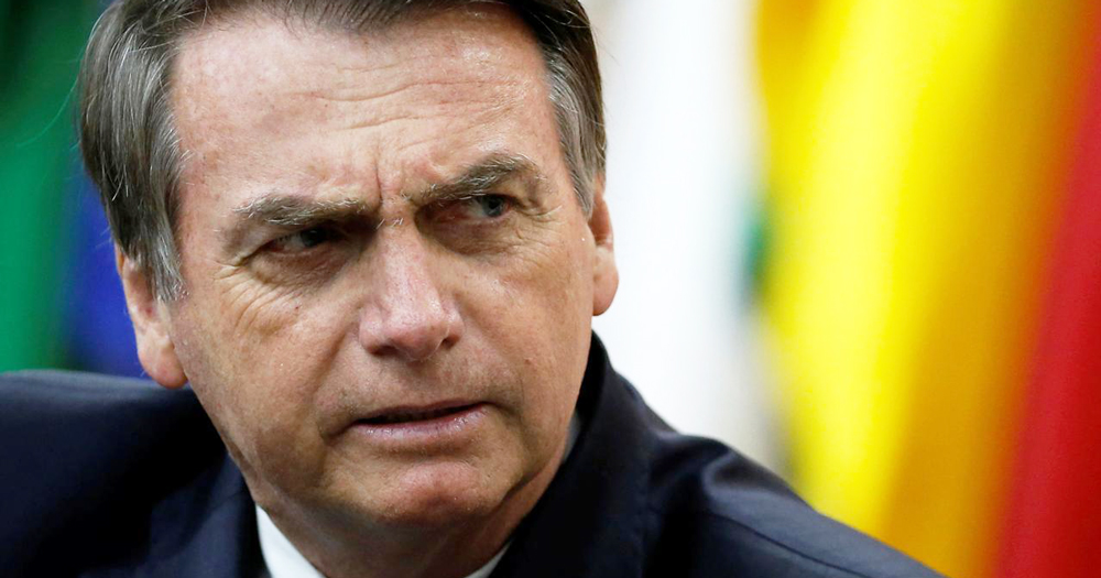 Brazilian president Jair Bolsonaro in a black suit in front of some flags, who wrote a Facebook post attacking the WHO.