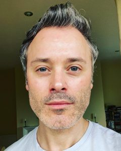 A man with grey stubble and slicked back hair takes a selfie