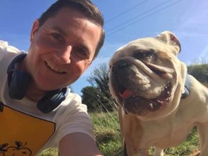 A smiling man in a t-shirt takes a selfie with a bulldog in a garden