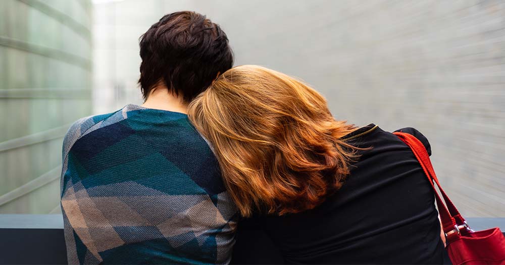 Seen from behind, a woman with long red hair rests her head on the shoulder of a woman with short dark hair