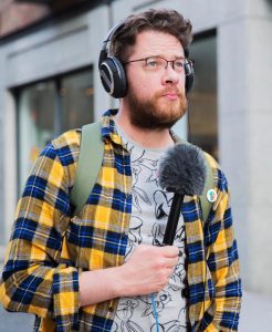 A bearded man stands on the street with headphones and a mic